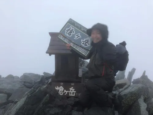 濃霧の槍ヶ岳山頂の祠の前で「槍ヶ岳3180m」の看板を掲げる登山者