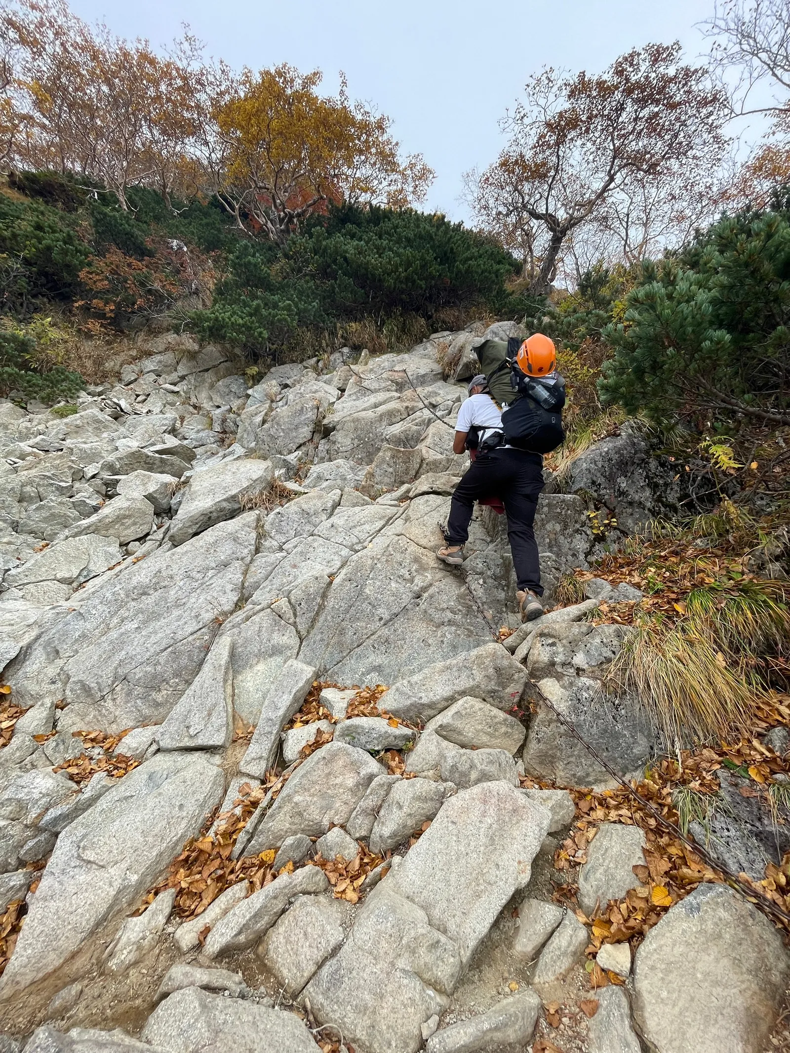 オレンジのヘルメットをかぶった登山者が、崩落地に近い岩のスラブ状の斜面を下っていく様子