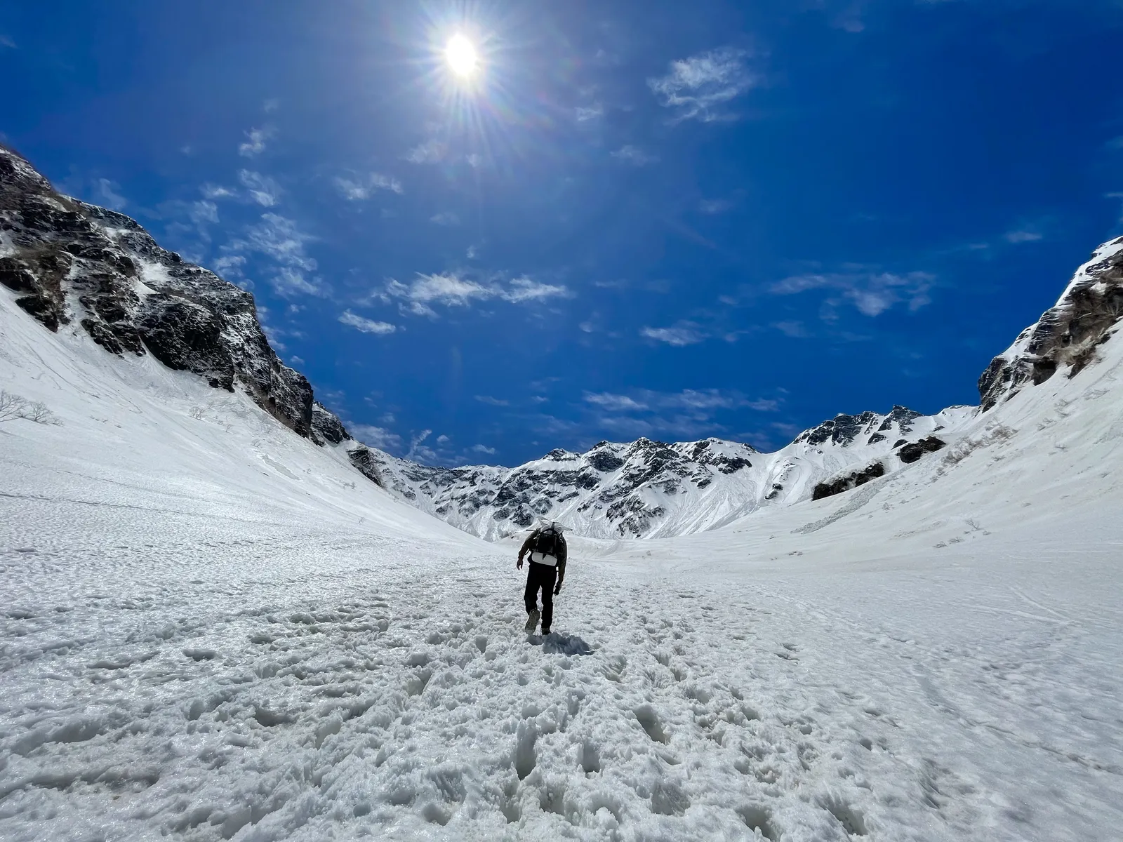 青空と太陽の下、両岸を雪壁に囲まれた広い雪の谷を登る登山者の後ろ姿