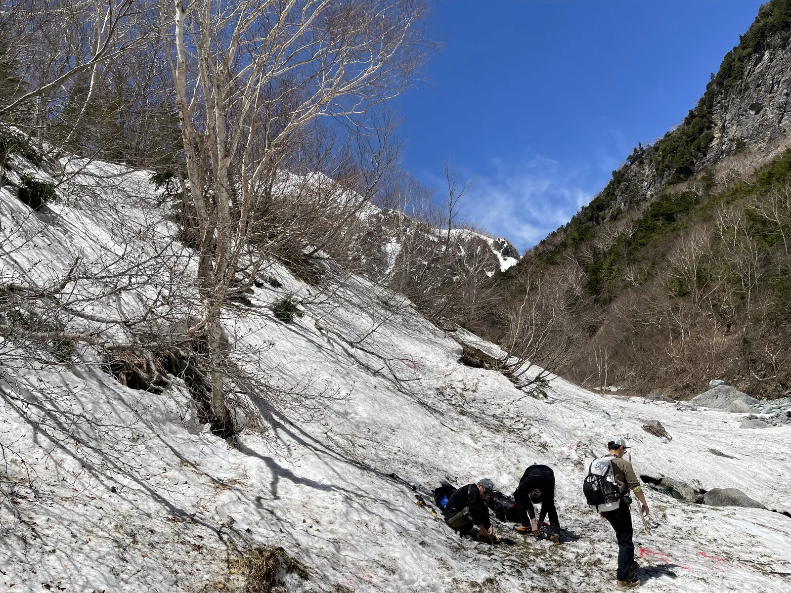 残雪期の雪渓の縁で、登山者たちがかがんで靴に滑り止めを装着している様子
